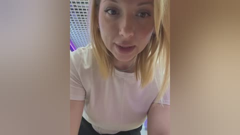 Media: Video of a fair-skinned, blonde woman in a white T-shirt, smiling, with a modern indoor background featuring a grid-patterned wall and purple lighting.