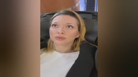 Media: Video of a Caucasian woman with straight, shoulder-length blonde hair, wearing a white blouse and black blazer, seated in a black leather chair, looking contemplative. Background includes a wooden wall and a window with closed blinds.