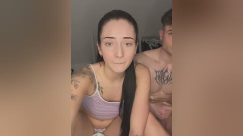 Media: Video of a fair-skinned woman with long black hair, wearing a striped crop top, sitting on a bed in a dimly lit room. She has tattoos on her arms and a tattooed man lies behind her.