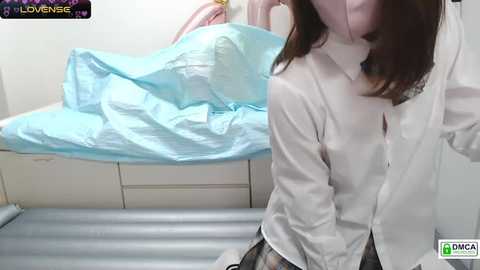 Media: A video of an Asian woman in a white button-up shirt, kneeling on a bed, with a pink mask and blue sheets in the background.