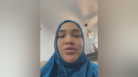 Media: Video of a young woman with light brown skin, wearing a blue hijab, sitting in a modern, dimly-lit room with a chandelier.