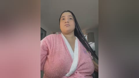 Media: Video of a plus-sized, dark-skinned woman with long braids wearing a pink, fuzzy robe, standing indoors with blurred background.