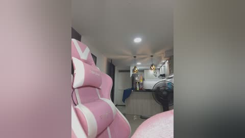 Media: A video of a modern gaming room featuring a pink gaming chair and a black fan in the foreground, with a minimalist kitchen and white appliances in the background.