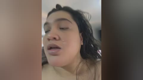 Media: Video of a woman with wet, dark hair, wearing minimal makeup, eyes closed, mouth slightly open, standing in a blurred bathroom setting.