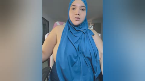 Media: Video of a plus-sized, light-skinned woman with a blue hijab and black pants, standing indoors with a neutral background.