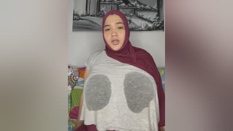 Media: A video of a young woman with a medium complexion, wearing a burgundy hijab and a gray T-shirt with large, gray, heart-shaped cutouts, seated against a white wall with a black-and-white landscape painting above her.