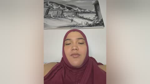 Media: Video of a smiling, light-skinned woman with brown hair and a maroon hijab, wearing a maroon robe, indoors with a black-and-white landscape print on a white wall behind her.