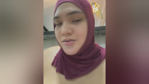 Media: Video of a young Asian woman with medium skin tone, wearing a maroon hijab, and a light-colored top, with blurred background showing a modern room.