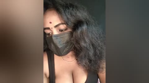 Media: Video of a woman with medium brown skin, wearing a black mask, black top, and a bindi. Her long, curly black hair partially covers her face. The background is dark.