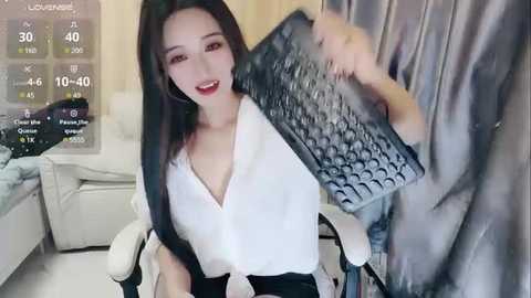 Media: A video of an East Asian woman with long black hair, wearing a white blouse, holding a grey patterned bag, seated on a white chair, in a modern living room.