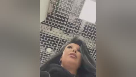 Media: A video of a woman with long, dark hair, possibly of Asian descent, wearing a black dress, standing in a tiled bathroom with a grid ceiling and fluorescent lights.