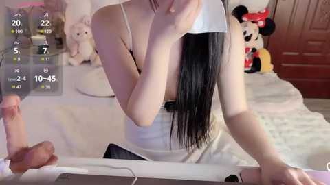 Media: A video of an East Asian woman with long black hair, wearing a white tank top, taking a mirror selfie in a bedroom. The room features a bed, plush toys, and a digital clock showing 20:22.