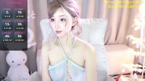 Media: Video of a young, fair-skinned Asian woman with shoulder-length blonde hair, wearing a light blue halter top, seated on a white chair in a softly lit room with a digital display showing her stats.