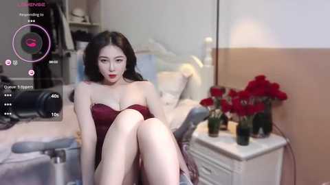 Media: Video of a young Asian woman with long black hair, wearing a strapless burgundy dress, sitting on a bed in a bedroom. Background includes a white nightstand with red roses and a mirror.