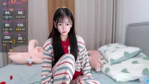 Media: A video of an Asian woman with long black hair and fair skin, wearing a red sweater and striped blanket, sitting on a bed with pastel pillows and a stuffed animal in a cozy room.