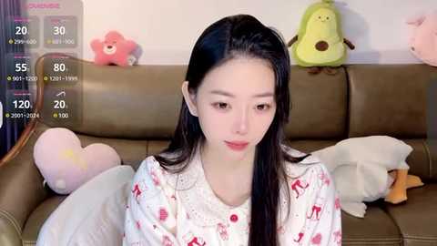 Media: Video of an East Asian woman with long black hair, wearing a red reindeer pajama top, sitting on a beige couch with stuffed animals, including a pink bear and green frog.