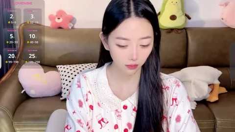 Media: A video of an East Asian woman with long black hair, wearing a white robe with red reindeer print, sitting on a brown leather couch. Background includes stuffed animals, a calendar, and a bookshelf.