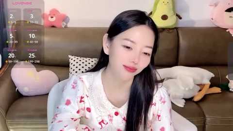 Media: A video of a young Asian woman with long black hair, wearing a white nightgown adorned with red hearts, sitting on a brown leather couch. Background features a calendar and stuffed toys, including a pink heart, a yellow frog, and a white duck.