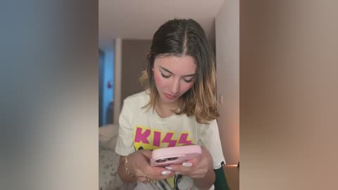 Media: A young woman with shoulder-length, ombre hair in a hallway, wearing a white \"KILL\" T-shirt, focused on her pink smartphone.