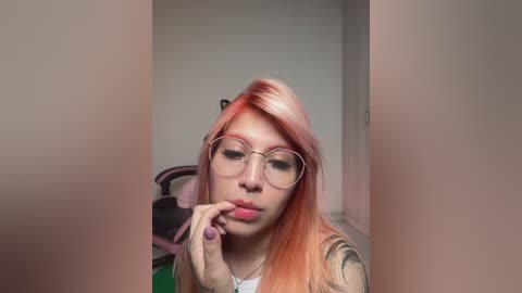 Media: Video of a young woman with light skin, wearing glasses, pink lipstick, and long, dyed orange hair, biting her finger, seated in a pink gaming chair.