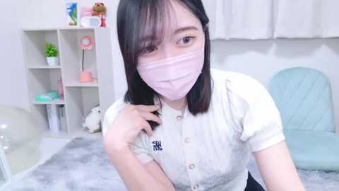 Video of an East Asian woman with straight black hair, wearing a pink face mask and a white button-up shirt, sitting on a plush light blue chair in a minimalist, white-themed room with a fluffy gray rug.