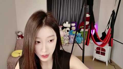 Media: A video of an East Asian woman with long, straight, dark hair, wearing a black sleeveless top, smiling. The background features a room with stuffed toys, a hammock, and a black curtain.