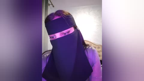 Media: A video of a person with long brown hair wearing a black niqab, revealing only their eyes, standing indoors with a white wall and a bed in the background.