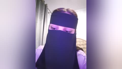 Media: Video of a woman in a black hijab covering her face, wearing a light-colored shirt, standing in a dimly lit room with gray curtains and a beige blanket.