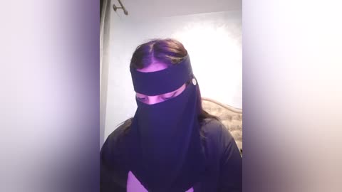 Media: A video of a person with long, dark hair, wearing a black face mask and black clothing, standing in a dimly lit room with a white wall and a bed visible in the background.