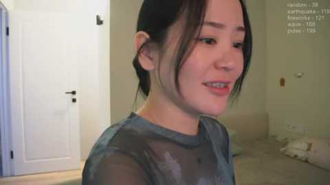 Media: A video of a young Asian woman with fair skin, dark hair pulled back, smiling. She wears a sheer, dark blue top. The background shows a minimalist, beige room with a door, a chair, and a white wall.