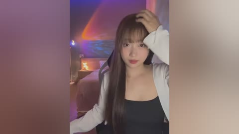 Media: Video of an Asian woman with long black hair, wearing a black top and white jacket, posing in a dimly lit room with colorful lights and a fireplace in the background.