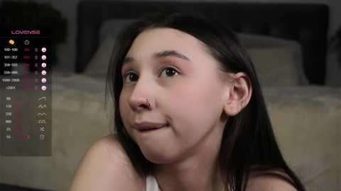 Media: Video of a young woman with long, straight black hair, light skin, and a nose ring, wearing a white tank top, sitting indoors. Calendar app with \"Loveme\" name visible in the background.