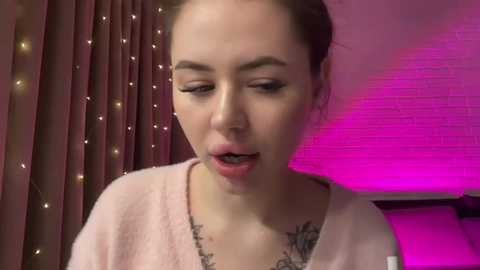 Media: Video of a young woman with fair skin, dark hair tied back, wearing a pink robe. She has a surprised expression with her mouth open, standing in a room with pink lighting and string lights on the left.