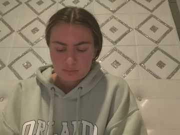 Media: Video of a young woman with fair skin, straight brown hair, and a neutral expression, wearing a green hoodie with \"USA\" on it. She sits on a tiled bathroom floor with a geometric pattern.