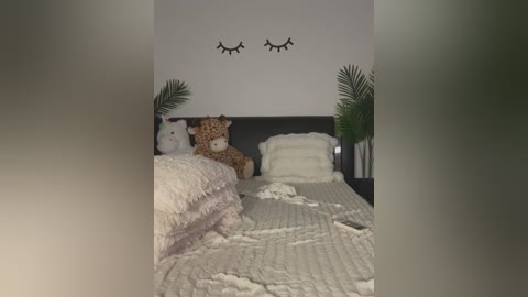 Media: Video of a cozy, dimly-lit bedroom with a dark wooden headboard. A plush giraffe toy and a white teddy bear are on a bed with a light, textured blanket. Black eyelash wall art and green palm plants flank the headboard.