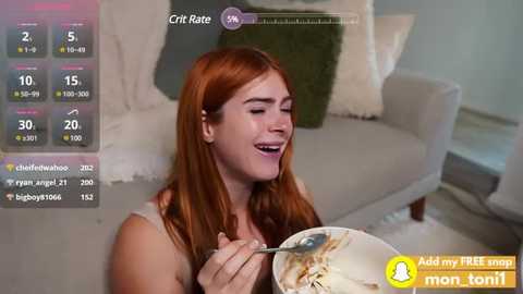 Media: Video of a fair-skinned woman with long, straight red hair, smiling, eating spaghetti, in a modern, light-colored living room with a white sofa and green throw pillows.