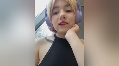 Media: Video of a fair-skinned young woman with short blonde hair, wearing large purple headphones, black halter top, and resting her chin on her hand, indoors with a blurred window background.