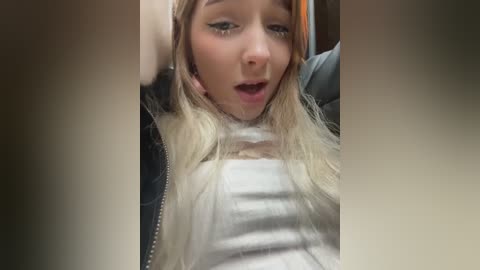 Media: Video of a blonde woman with fair skin, wearing a white striped shirt and black jacket, looking surprised and slightly open-mouthed.