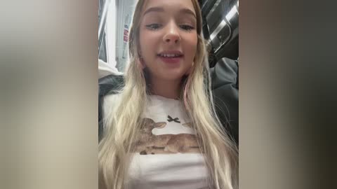 Media: Video of a young, fair-skinned woman with long, blonde hair, smiling slightly, wearing a white t-shirt with a graphic design. She sits in a dimly lit, narrow subway car, with blurred background details.