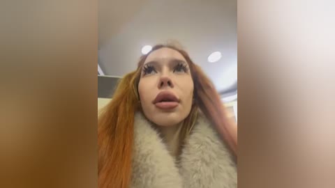 Media: Video of a woman with long, straight, orange hair, fair skin, and full, pink lips. She wears a white fur coat and looks upward, possibly indoors with blurred ceiling lights.