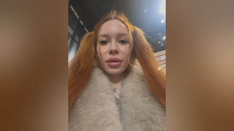 Media: A video of a young Caucasian woman with long, bright orange hair styled in pigtails, wearing a fur-trimmed coat, smiling. The background is blurred, suggesting an indoor setting with warm lighting.