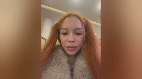 Media: Video of a young woman with long, vibrant orange hair styled in pigtails. She has a fair complexion, wearing a grey, fluffy jacket. The background is a dimly lit indoor setting with a white ceiling and beige walls.