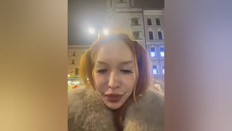 Media: Video of a young Asian woman with fair skin and red hair styled in pigtails, wearing a fur coat, making a silly face. Background shows a blurred urban scene at night with lit buildings and streetlights.