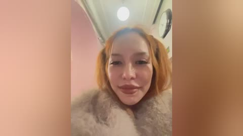 Media: Video of a young Asian woman with short, straight, orange hair, wearing a fur coat, smiling. The background features a pink wall and a ceiling light.