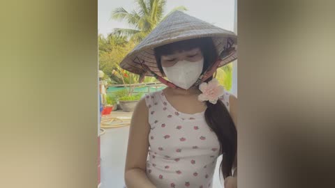 Media: Video of an Asian woman with long black hair, wearing a conical hat, white face mask, and a sleeveless floral dress, standing outdoors on a balcony with a palm tree in the background.