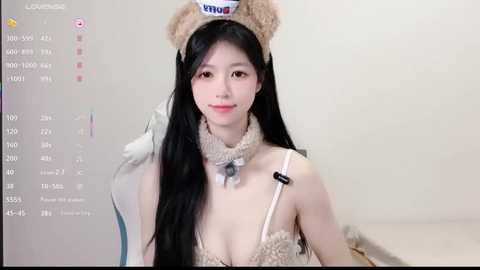 Media: Video of a young Asian woman with long black hair, wearing a teddy bear headband, beige lace dress, and a furry collar. Background shows a computer screen displaying a virtual reality interface.