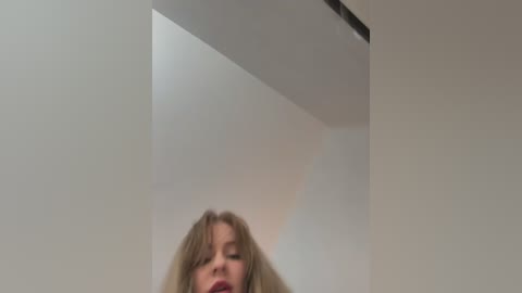 Media: Video of a blonde woman with long hair and closed eyes, standing in a dimly lit room with white walls and a ceiling light.