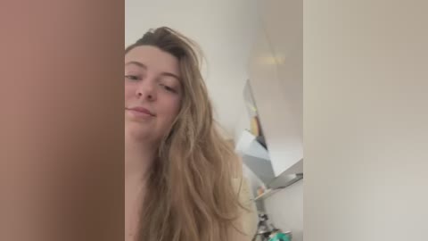 Media: A video of a young woman with long, wavy, light brown hair, fair skin, and a relaxed expression, partially obscured by a person's arm on the left. The background shows a modern kitchen with white cabinets and stainless steel appliances.