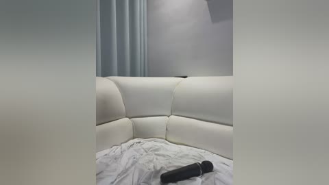 Media: Video of a modern, white, upholstered, curved sectional sofa with a minimalist, light gray wall and curtains in the background. A black vibrator is placed on the sofa's surface.