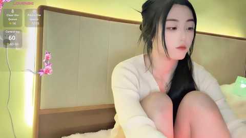 Media: Video of a young Asian woman with long black hair, sitting on a bed, wearing a white cardigan and black top, looking pensive. The room has a beige headboard and pink flowers on the wall.